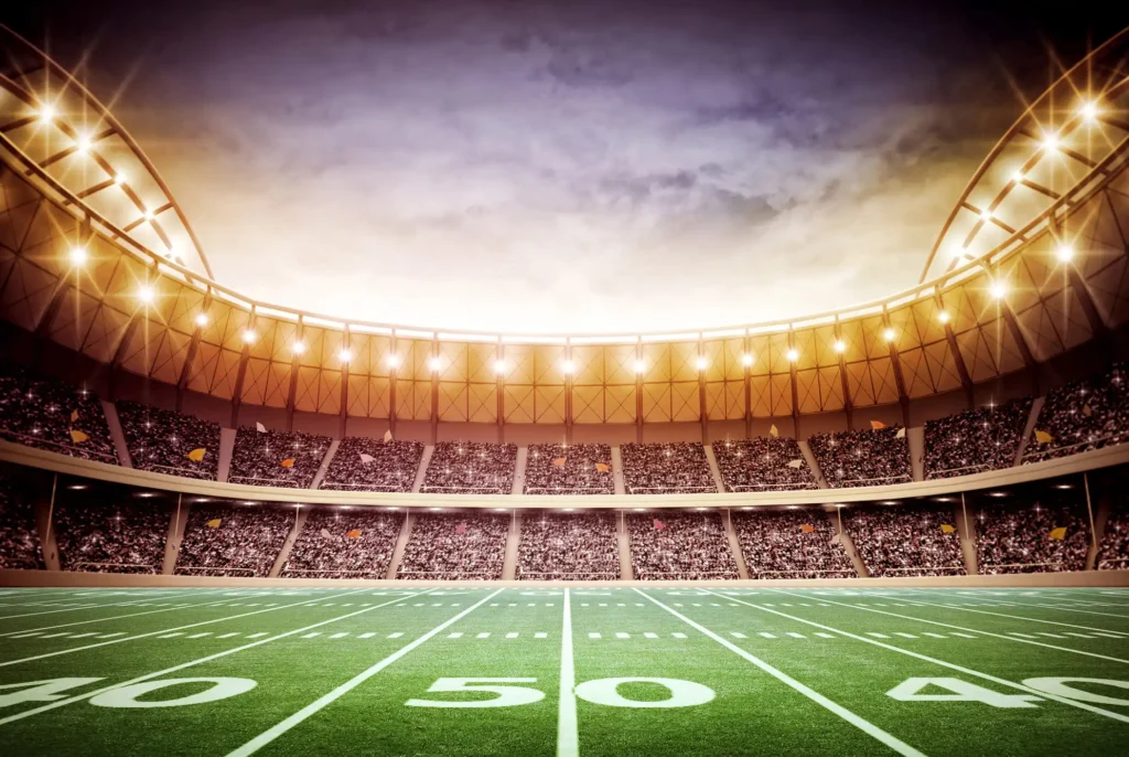 A large, brightly lit football stadium filled with spectators, viewed from the middle of the field, showing the 50-yard line under a dramatic sunset sky.