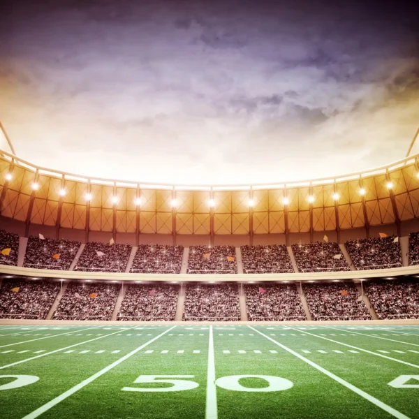 A large, brightly lit football stadium filled with spectators, viewed from the middle of the field, showing the 50-yard line under a dramatic sunset sky.