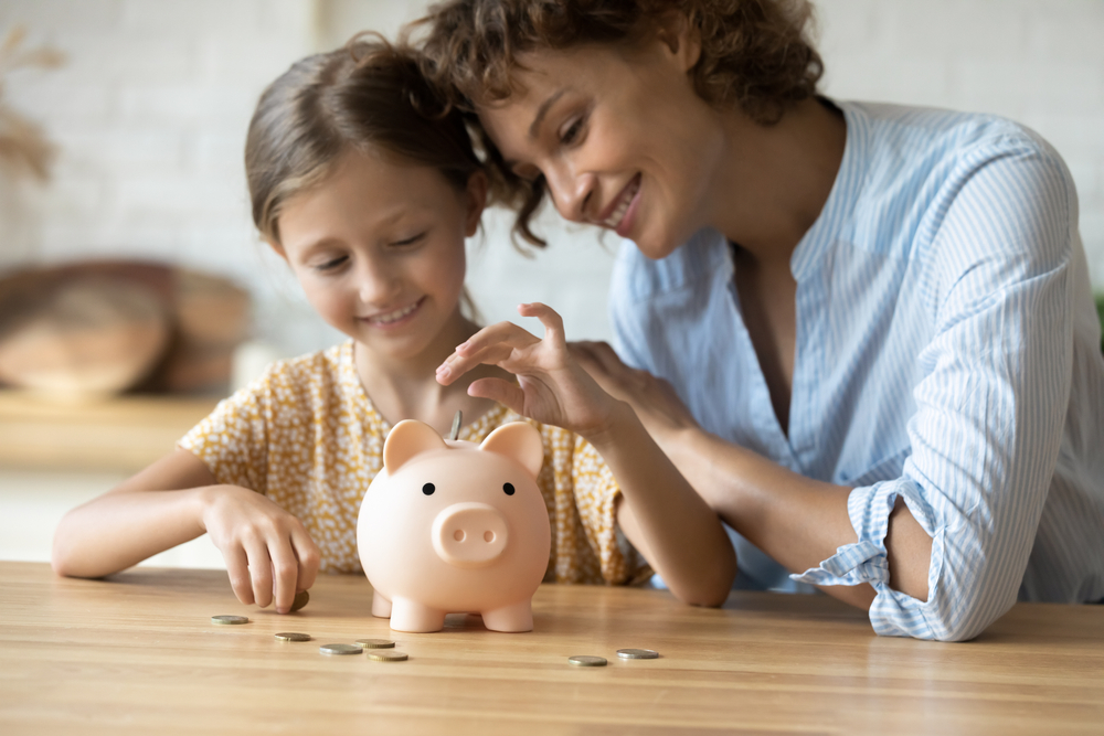A woman and a young girl smile as they put coins into a piggy bank on a wooden table, symbolizing saving money and financial education.