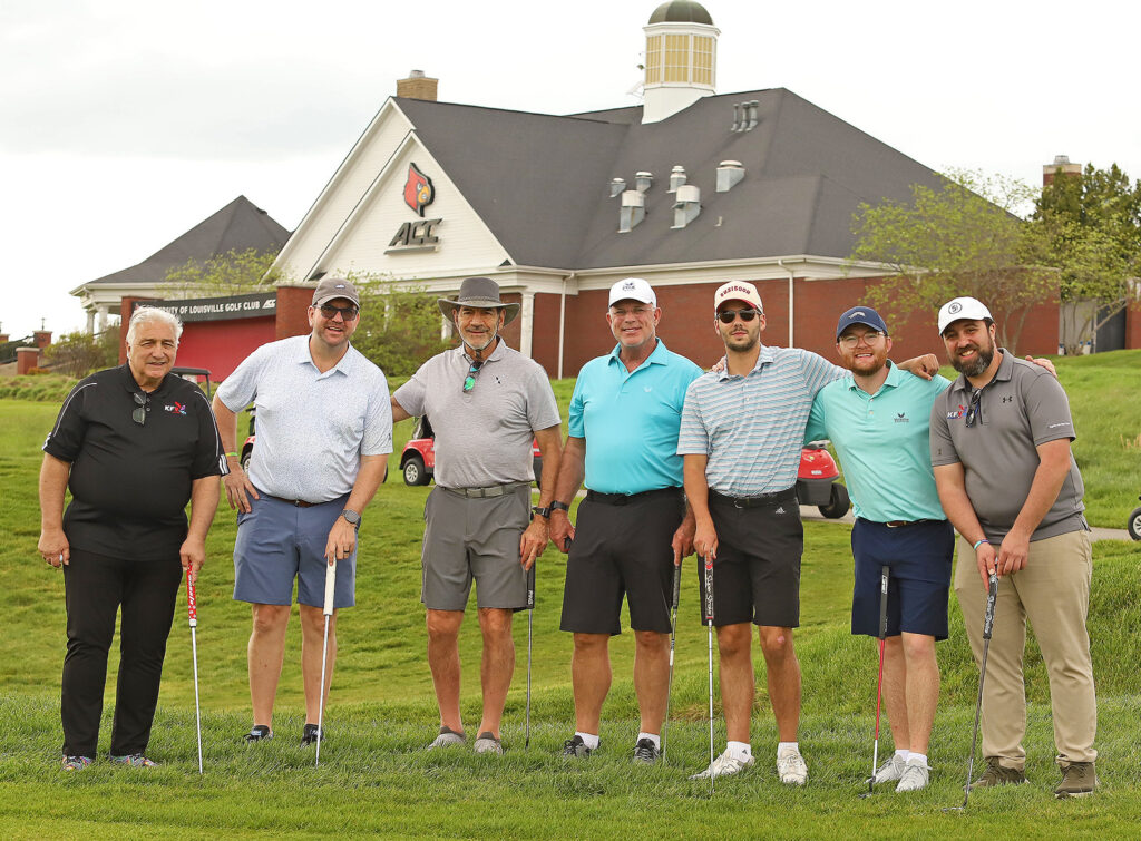 Seven men pose together on a golf course, each holding a golf club, with a building featuring the ACC and Louisville Cardinals logos in the background. They are dressed in casual golf attire and smiling.