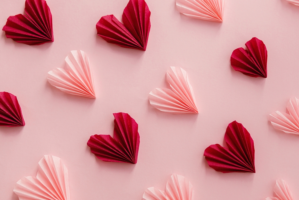 Pink and red origami paper hearts are arranged in a neat, diagonal pattern on a soft pink background, creating a romantic and decorative look.