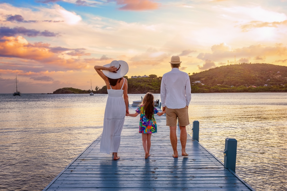 A family of three—woman, man, and young girl—walks hand in hand on a wooden dock toward the water at sunset. The sky is colorful, and hills and boats are visible in the distance.