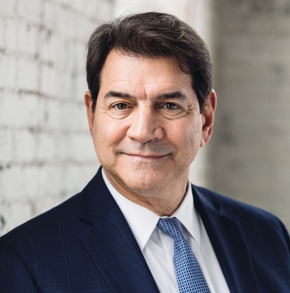 A middle-aged man with dark hair wearing a navy suit, white shirt, and light blue tie smiles slightly while standing in front of a white brick wall.