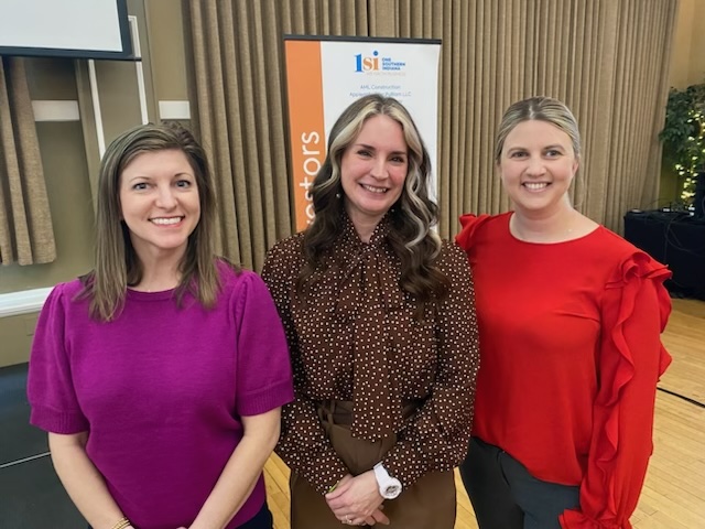 Three women stand indoors, smiling at the camera. The woman on the left wears a purple top, the center woman wears a brown polka dot blouse, and the woman on the right wears a bright red top. A presentation banner is seen in the background.