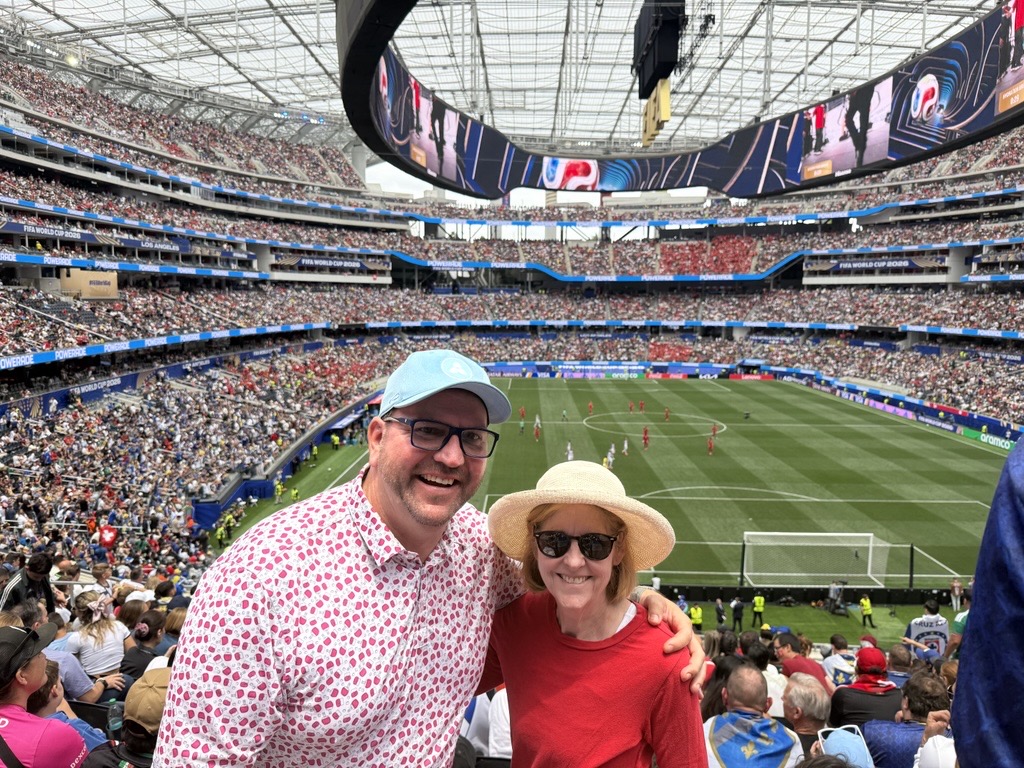 Two smiling people pose in front of a large, crowded soccer stadium with a match in progress. The man wears a blue cap and patterned shirt; the woman wears sunglasses, a straw hat, and a red shirt.