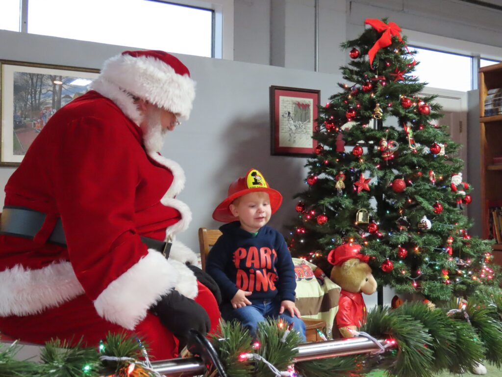 A child wearing a firefighter hat sits next to Santa Claus by a decorated Christmas tree indoors. The child is smiling, and the scene is festive with ornaments and greenery.