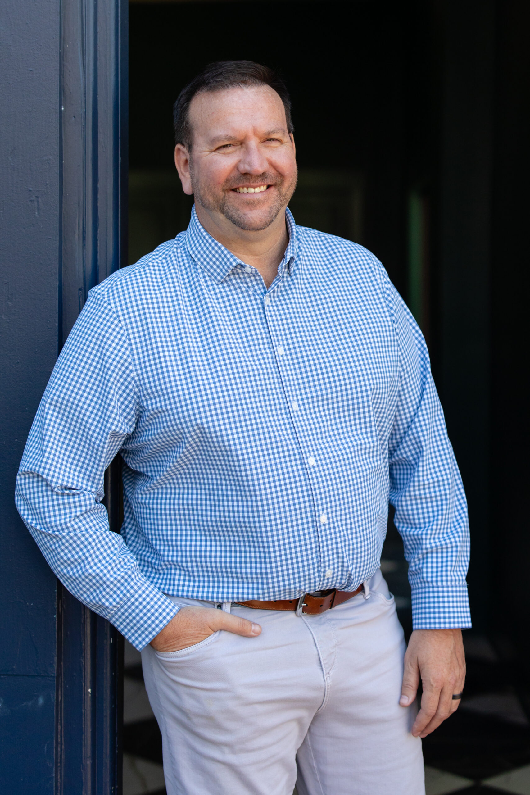 A man in a blue checkered shirt and light pants stands smiling with one hand in his pocket, leaning against a blue wall—a confident pose that echoes the professionalism of Vaughan Scott from Axiom FSG.