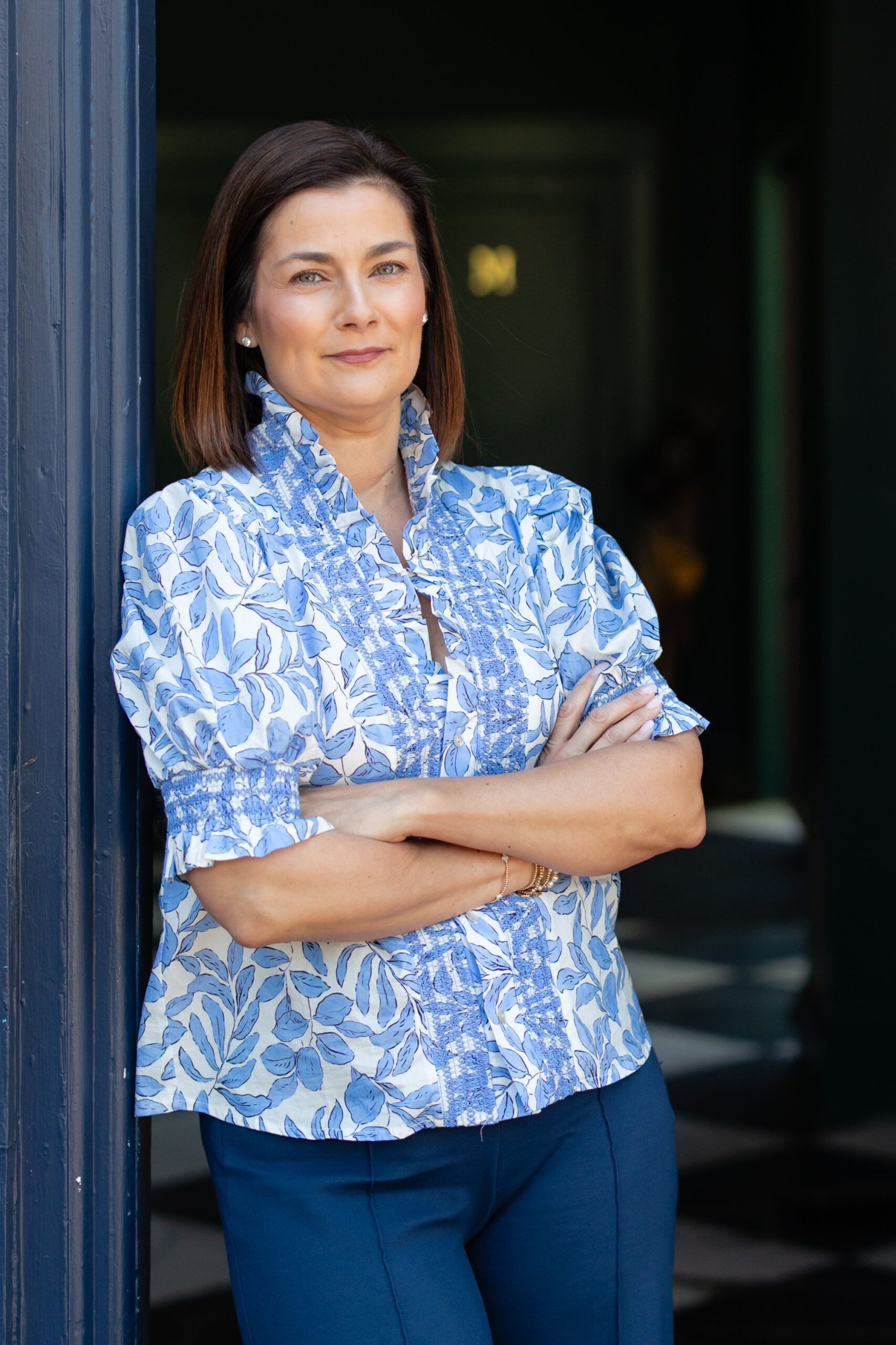 A woman with straight brown hair stands indoors, leaning against a doorframe with her arms crossed, wearing a blue and white patterned blouse and dark blue pants, and looking confidently at the camera—an image that perfectly captures the poised professionalism often associated with Abby Wolfe Axiom FSG.