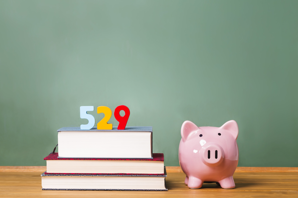 A pink piggy bank sits beside a stack of books topped with colorful numbers 529 in front of a green chalkboard, symbolizing education savings.
