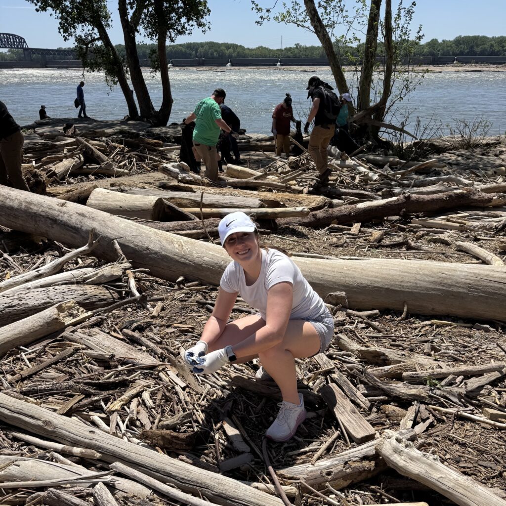 A woman in a white cap and gloves crouches and smiles while picking up debris on a riverbank covered with driftwood; other people clean up in the background near the water.