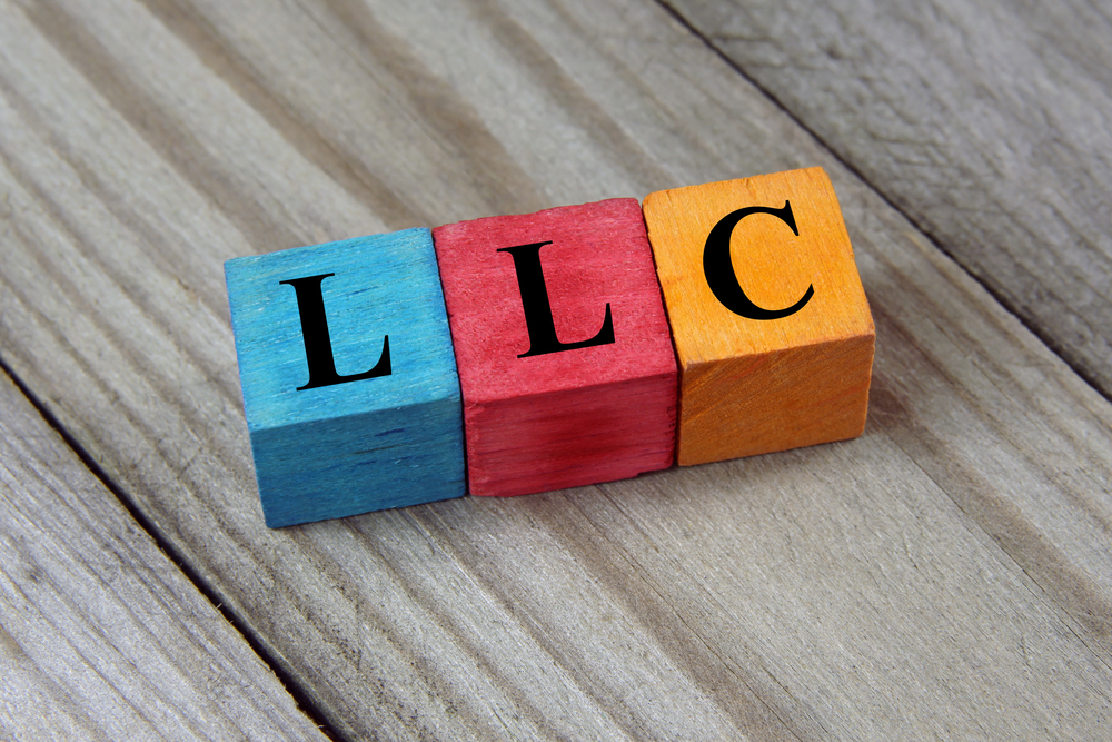 Three wooden blocks on a wooden surface, each painted a different color (blue, red, orange) and each displaying one black letter to spell LLC.