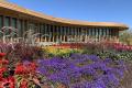 A modern building with large vertical windows sits behind vibrant flower beds filled with purple, red, and yellow blooms under a clear blue sky.