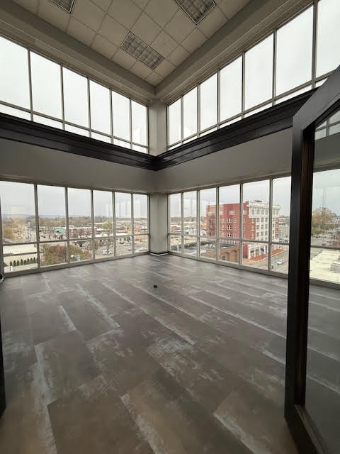 A spacious empty corner office with large floor-to-ceiling windows on two sides, offering a panoramic city view and ample natural light, with gray flooring and high ceilings.