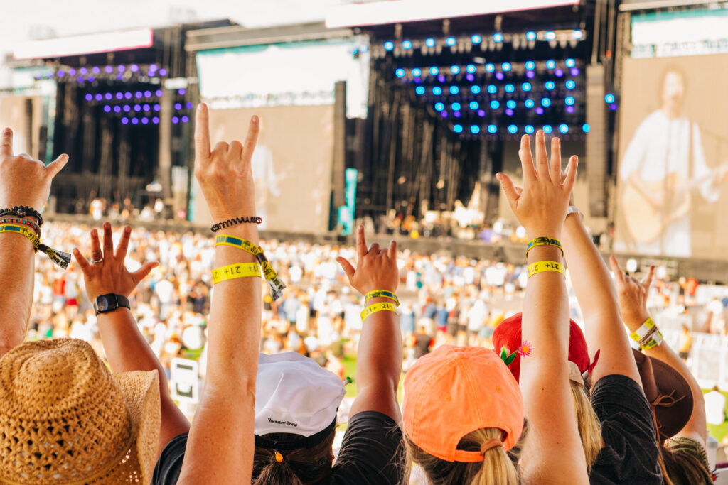 People at an outdoor concert raise their hands toward the stage, wearing yellow wristbands. The stage is brightly lit with blue lights and a large crowd is visible in the background.