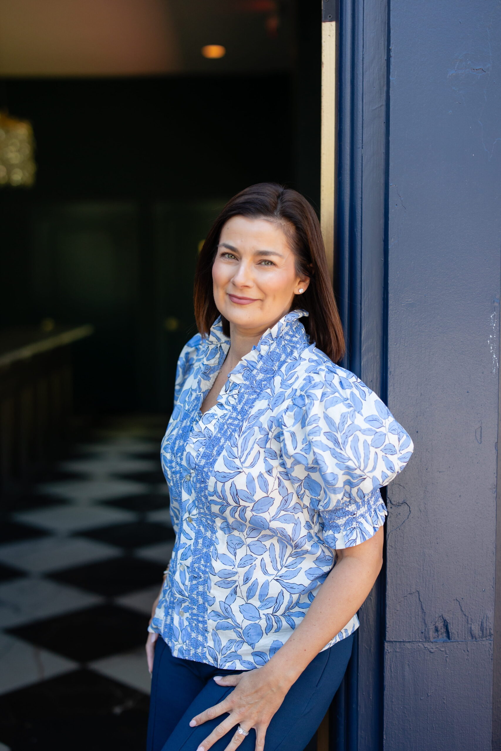A woman with straight brown hair stands, smiling, in front of a dark blue wall—her confident demeanor suggesting the professionalism you’d expect from a certified financial planner near me. She wears a blue and white patterned blouse and navy pants, while the floor inside features a striking black and white checkered pattern.
