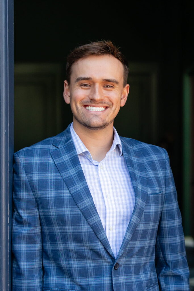 A smiling man wearing a blue plaid suit jacket and a light checkered shirt stands by a doorway with a dark background, embodying the approachable professionalism you’d expect from a certified financial planner near me.