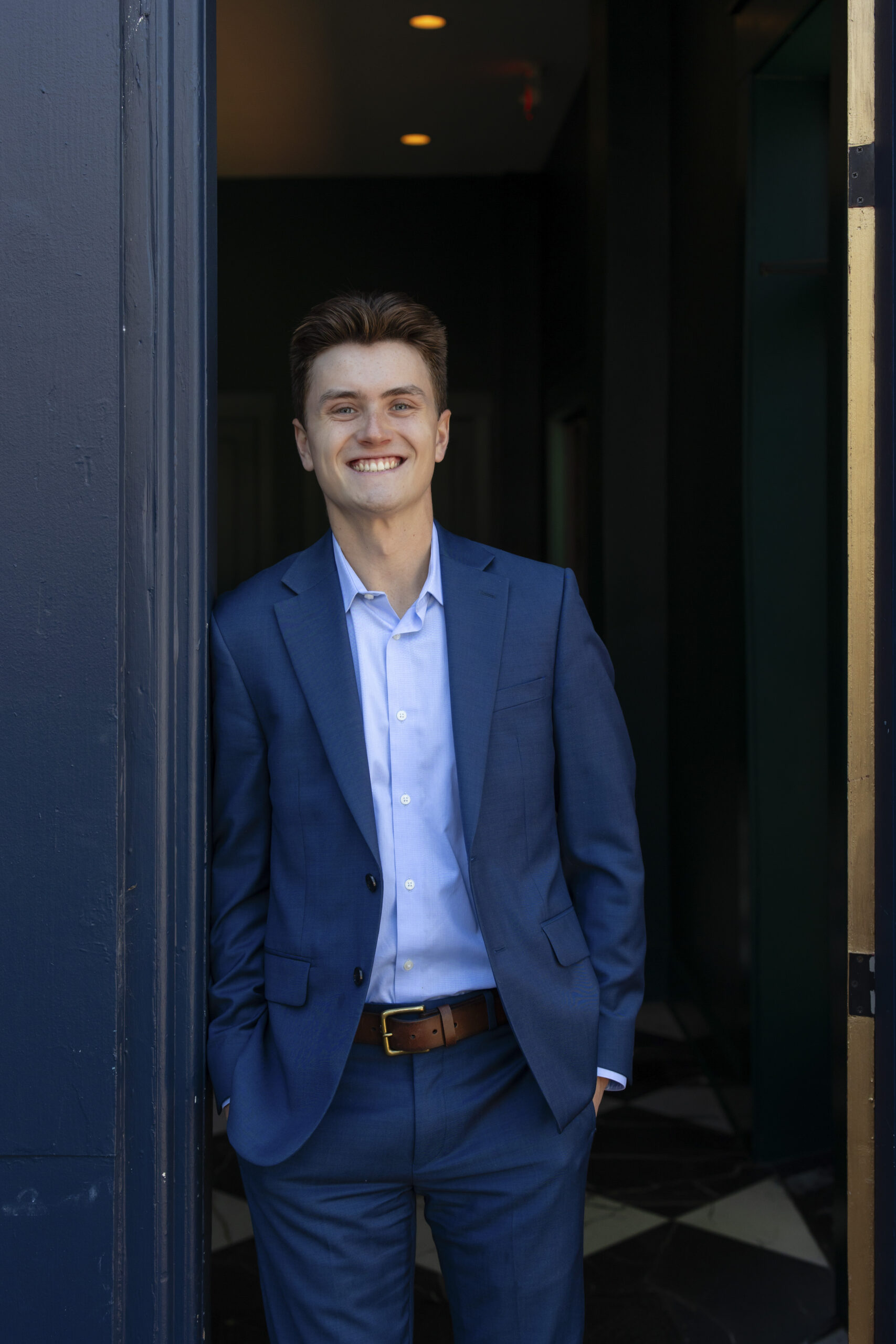 A young man, Garrett Huber, in a blue suit and light blue shirt stands smiling in a doorway, with one hand in his pocket, against a dark background with soft lighting.
