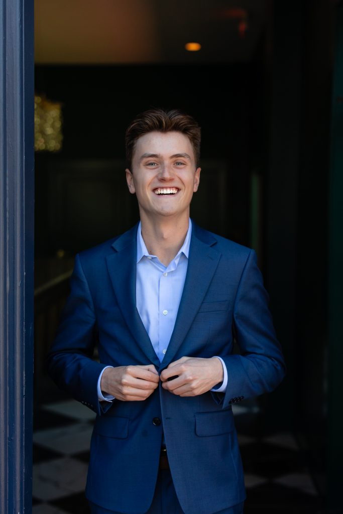 A young man in a blue suit, perhaps a certified financial planner near me, smiles while buttoning his jacket, standing in a doorway with a dark interior behind him.