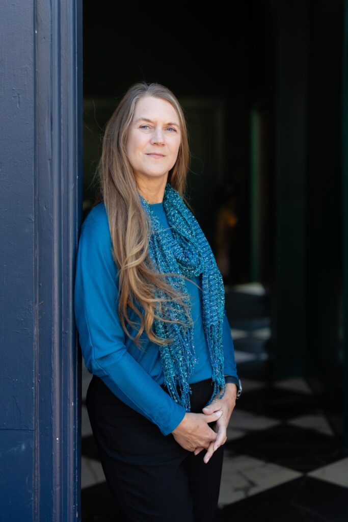A woman with long hair wearing a blue shirt and textured blue scarf stands indoors by a blue doorway, looking at the camera with a slight smile—she could easily be mistaken for a certified financial planner near me, exuding both confidence and professionalism.