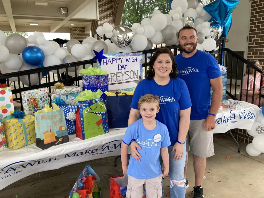A smiling family stands in front of a table filled with colorful presents and a HAPPY WISH DAY REYSON sign at a Make-A-Wish event, surrounded by white and blue balloons and decorations.