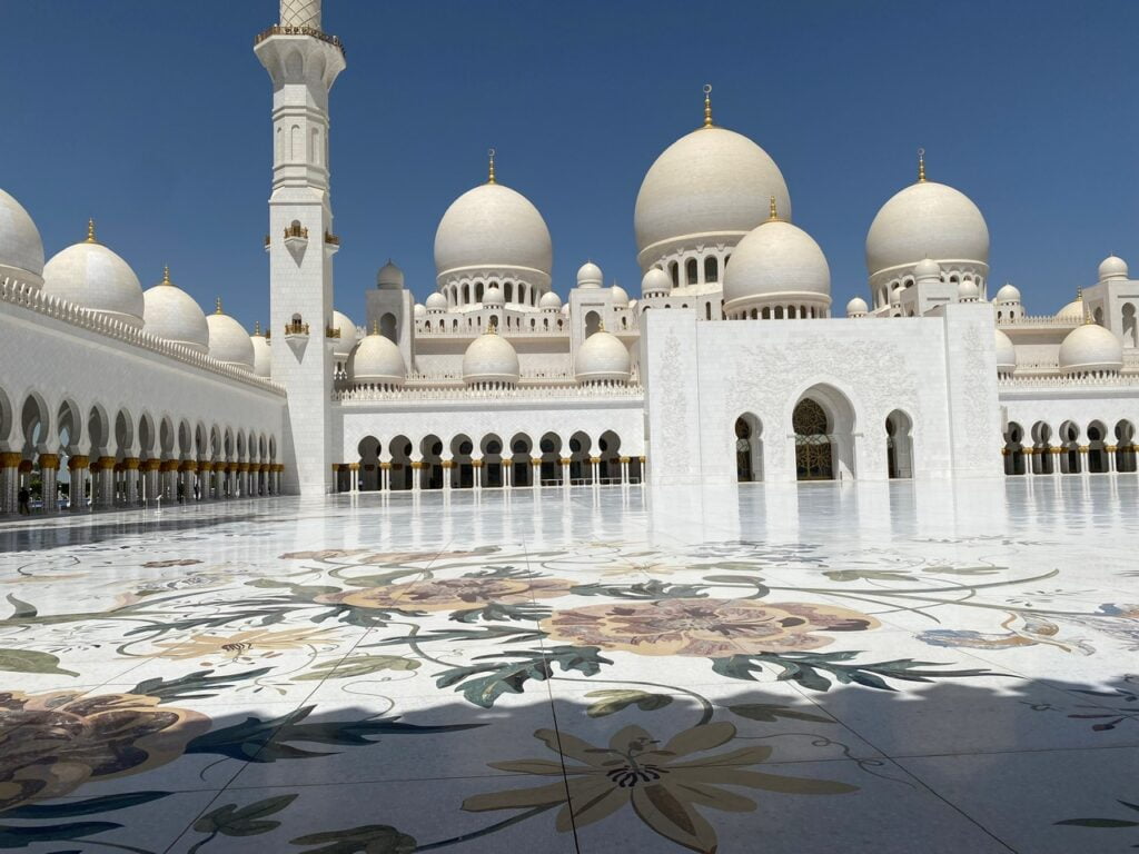A bright, sunny view of the Sheikh Zayed Grand Mosque with white domes, arched entrances, and a spacious courtyard featuring detailed floral designs on the polished marble floor.