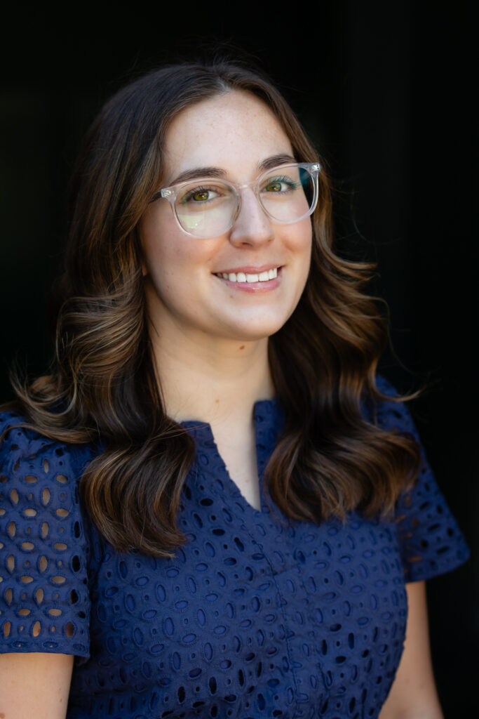 A woman with long brown wavy hair, wearing clear glasses and a navy blue eyelet dress, smiles in front of a dark background—her confident demeanor reflecting the professionalism of Jac Scott Fitzpatrick FPQP.