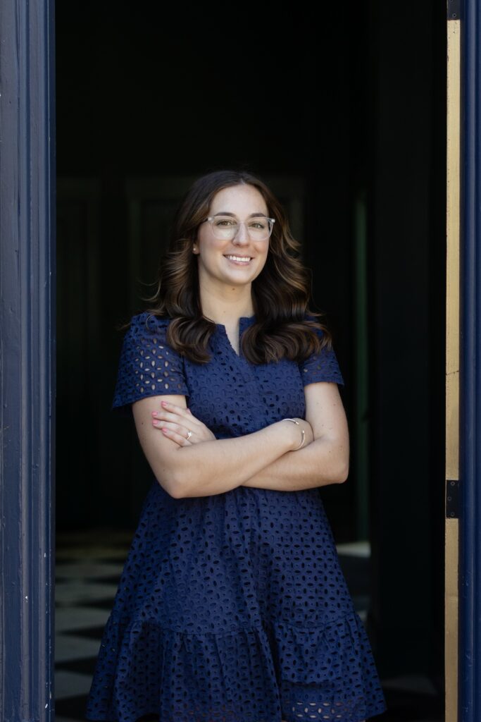 A woman with long brown hair, glasses, and a navy blue dress stands in a doorway with her arms crossed, smiling confidently at the camera—exuding the approachable professionalism you’d expect from a certified financial planner near me.