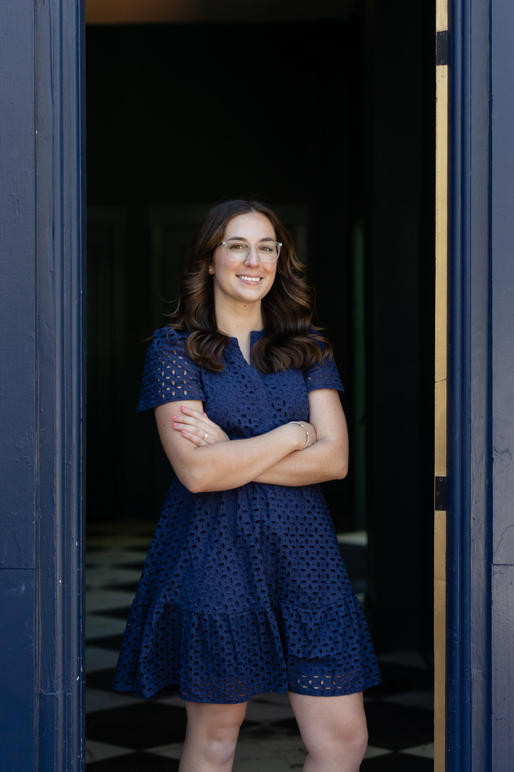 Jac Scott Fitzpatrick FPQP stands smiling with arms crossed in a doorway with dark blue walls and a checkered floor inside. She is a woman with long brown hair and glasses, wearing a short-sleeved navy dress.