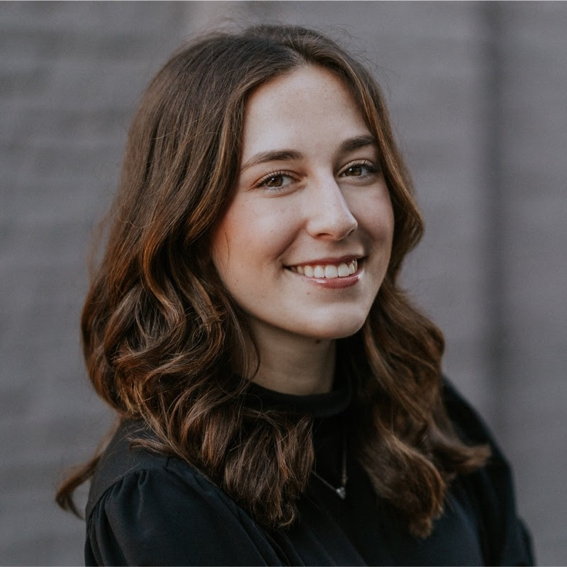 A young woman with wavy brown hair and a black top smiles warmly at the camera, standing in front of a blurred gray brick wall.