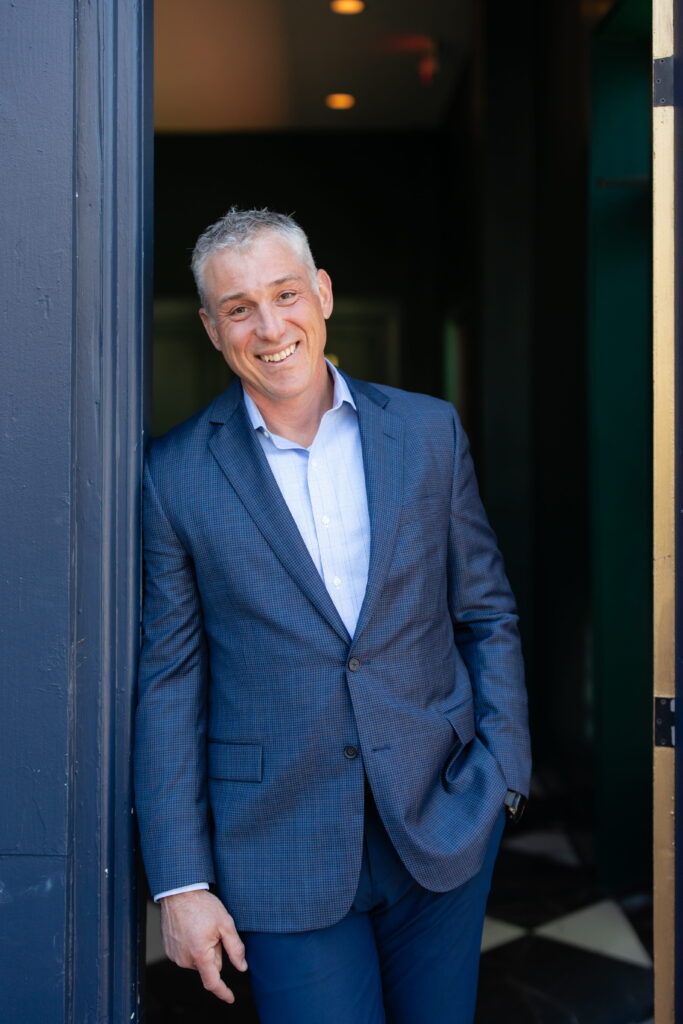 Smiling man with short gray hair in a blue suit leans casually against a navy wall indoors, exuding confidence and professionalism—just the demeanor you’d expect from a certified financial planner near me—with one hand in his pocket and the other resting by his side.