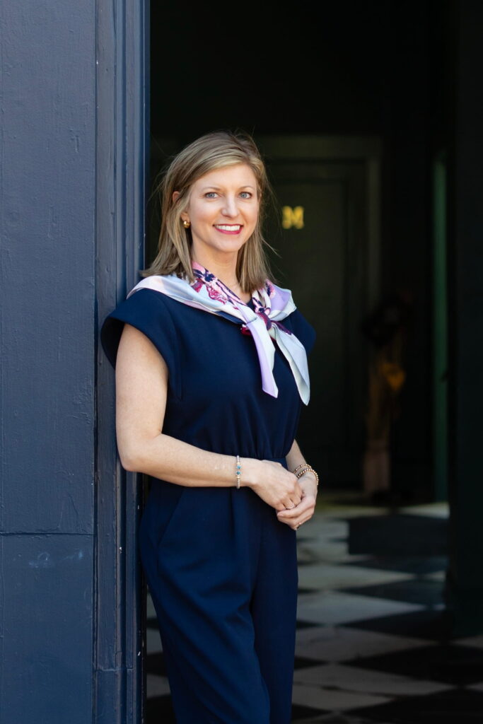 A woman in a navy outfit with a patterned scarf stands by a blue wall, smiling confidently, evoking the approachable professionalism you’d expect from a certified financial planner near me. Behind her, a dark, stylish interior and checkerboard floor complete the sophisticated atmosphere.