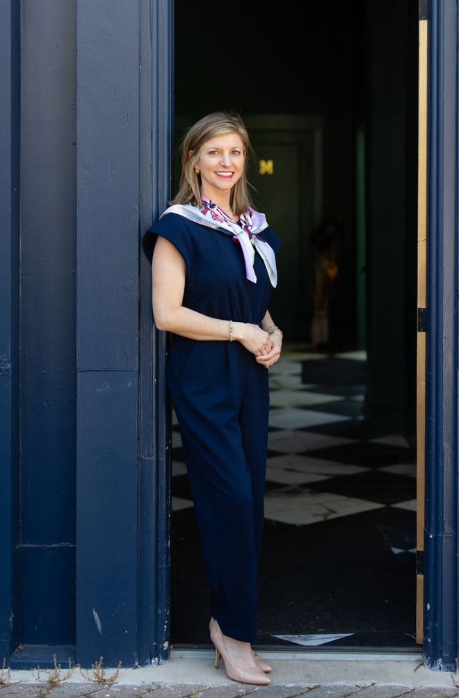 Leah Driver CPA, dressed in a navy jumpsuit and pink heels, stands in a doorway, smiling. She wears a light-colored scarf around her neck, while the floor inside features a striking black-and-white diamond pattern.
