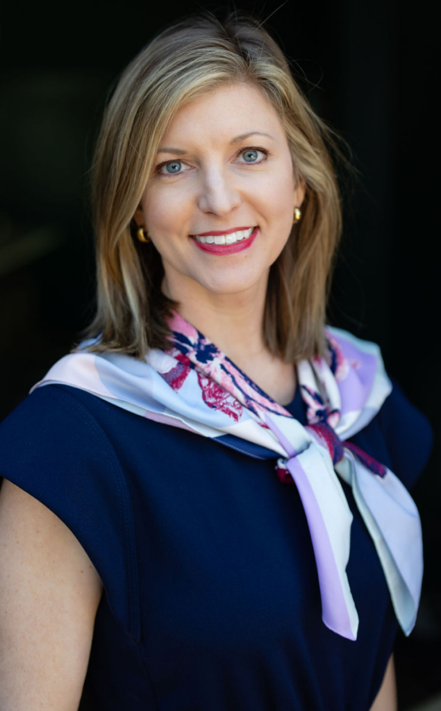 A woman with shoulder-length blonde hair, wearing a navy dress and a light-colored scarf with floral patterns, smiles at the camera against a dark background. Leah Driver CPA exudes both professionalism and warmth in this poised portrait.