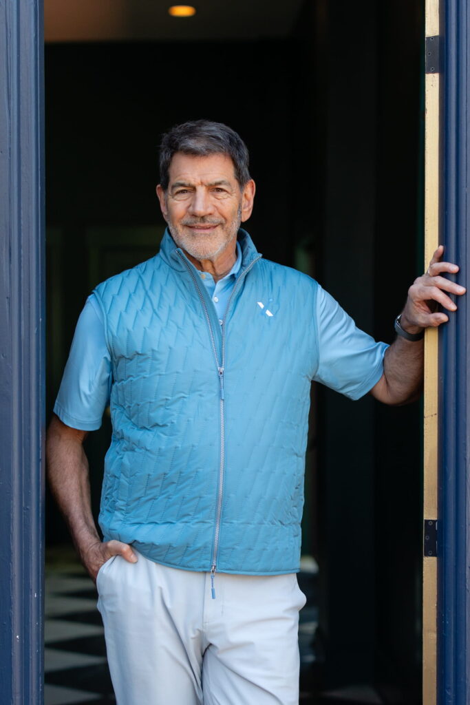 A middle-aged man with short dark hair and a beard stands in a doorway, wearing a light blue quilted vest over a pale blue shirt and white pants, smiling softly with one hand resting on the open doorframe—resembling the approachable confidence you might expect from a certified financial planner near me.
