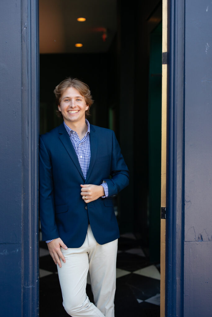 A person wearing a navy blazer, checkered shirt, and light pants stands in a doorway, smiling and holding their jacket lapel with one leg bent—exuding the approachable confidence of a certified financial planner near me.