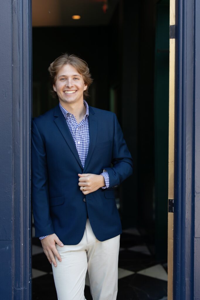 A young man wearing a navy blue blazer, checkered shirt, and light pants stands smiling in a doorway, adjusting his jacket with one hand—looking every bit the approachable professional you’d expect from a certified financial planner near me.