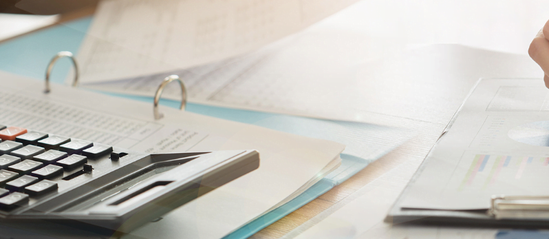 A close-up of a calculator, printed documents, a binder, and a hand holding a pen, suggesting someone working on financial calculations or office paperwork at a desk.