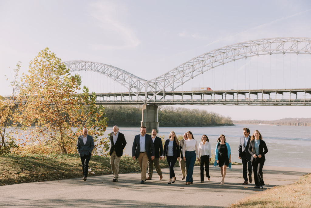 A group of people in business attire, possibly searching for a certified financial planner near me, walk together on a paved path by a river, with a large arched bridge and autumn trees in the background under a blue sky.