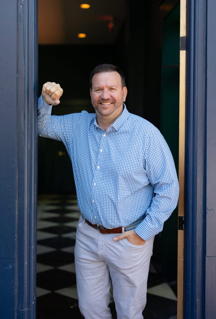 A man in a blue checkered shirt and light pants stands in a doorway, smiling and resting his elbow on the doorframe. The background has a black and white checkered floor and dark walls, creating an inviting atmosphere reminiscent of meeting with a certified financial planner near me.