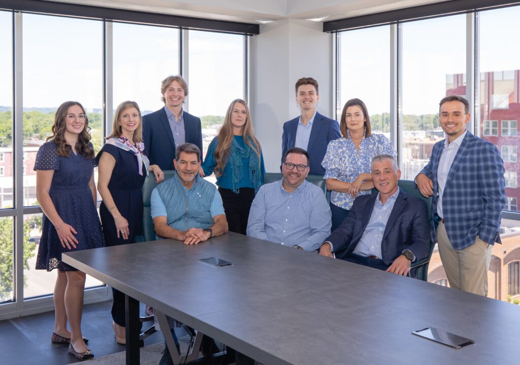 Whole Team Conference Room - Cropped Ten professionally dressed people, six standing and four seated, pose for a group photo in a modern office with large windows and a cityscape view in the background, reflecting the collaborative spirit at the core of About Axiom Financial.