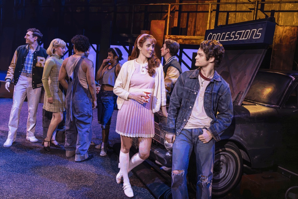 A group of young adults in retro clothing stand near a car and concessions sign on stage, two are talking up front while others gather near the car in the background.