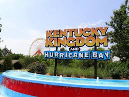 A large sign for Kentucky Kingdom and Hurricane Bay stands in front of a blue water feature, with greenery and a Ferris wheel visible in the background under a clear sky.