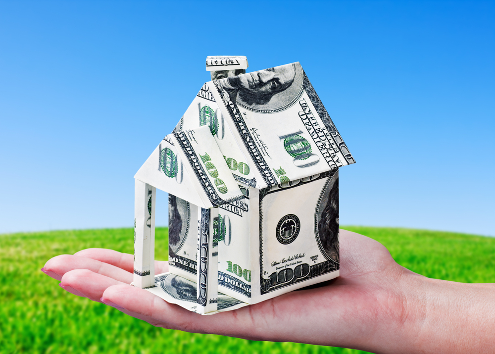 A hand holds a small house made of folded US hundred-dollar bills, set against a grassy field and clear blue sky.