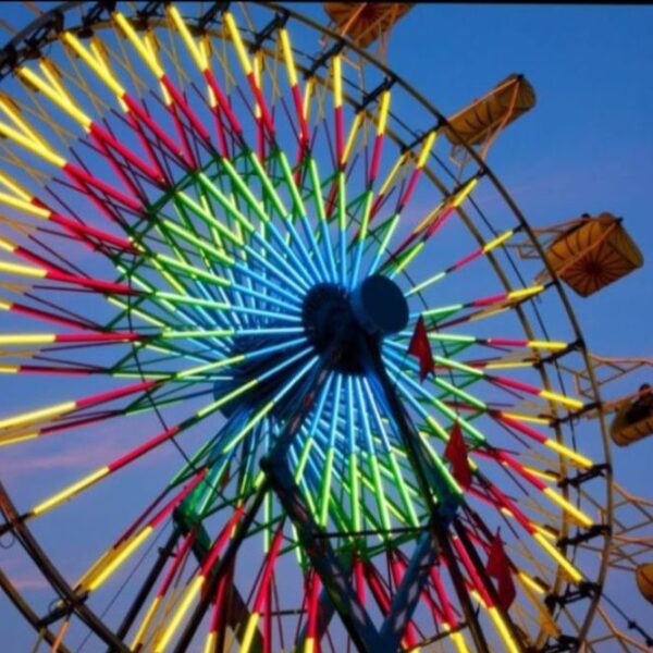 A brightly lit Ferris wheel glows with neon yellow, blue, green, and red lights against a pink and blue evening sky. Yellow passenger cabins are visible along the wheel.