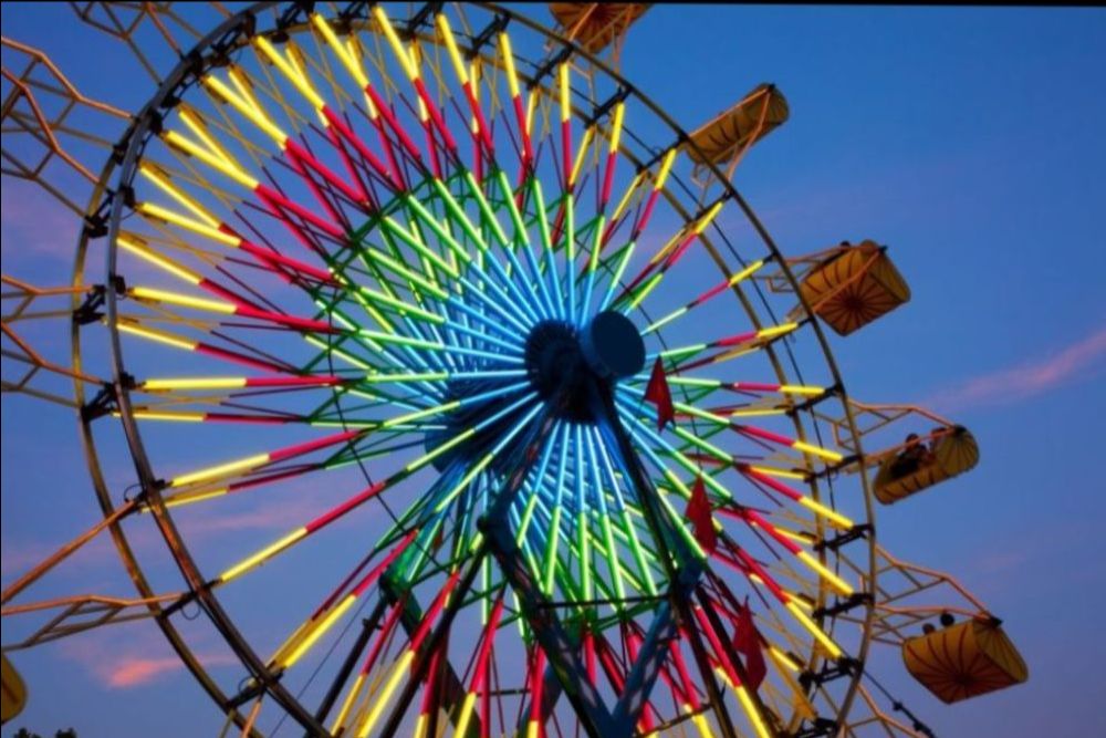 A brightly lit Ferris wheel glows with neon yellow, blue, green, and red lights against a pink and blue evening sky. Yellow passenger cabins are visible along the wheel.