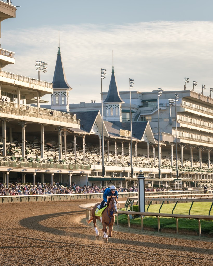 A jockey riding a racehorse on a dirt track at Churchill Downs, with the iconic twin spires and grandstands in the background under a clear sky.