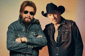 Two men pose together against a neutral background. One wears sunglasses, a denim jacket, and has a beard with arms crossed. The other wears a black cowboy hat and a dark shirt with a few buttons undone.