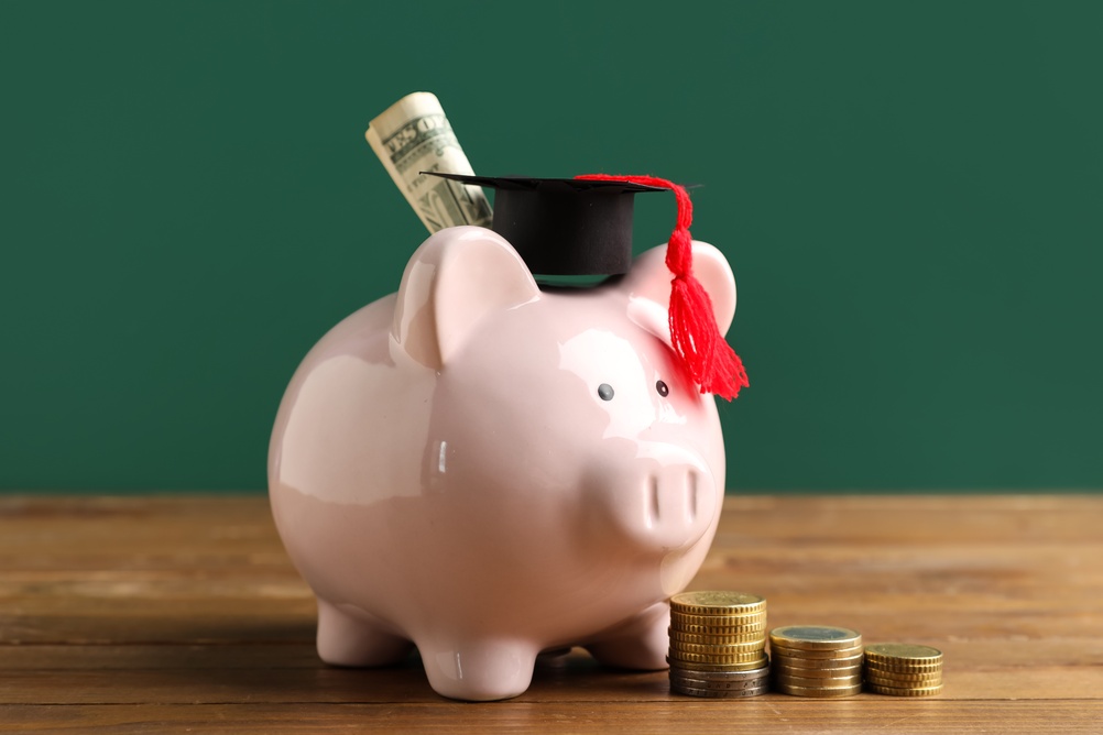 A pink piggy bank with a graduation cap and a rolled dollar bill inside, next to stacks of coins, on a wooden surface with a green background.