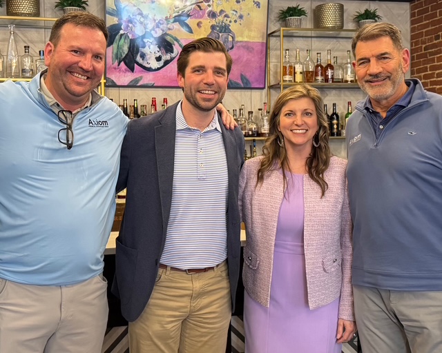 Four people stand together smiling in front of a bar with shelves of bottles and a colorful floral painting on the wall. The group includes three men and one woman, all dressed in business casual attire.