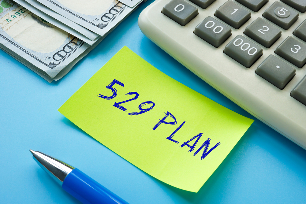 A yellow sticky note with 529 PLAN written on it sits next to a stack of hundred-dollar bills, a calculator, and a blue pen on a blue background.
